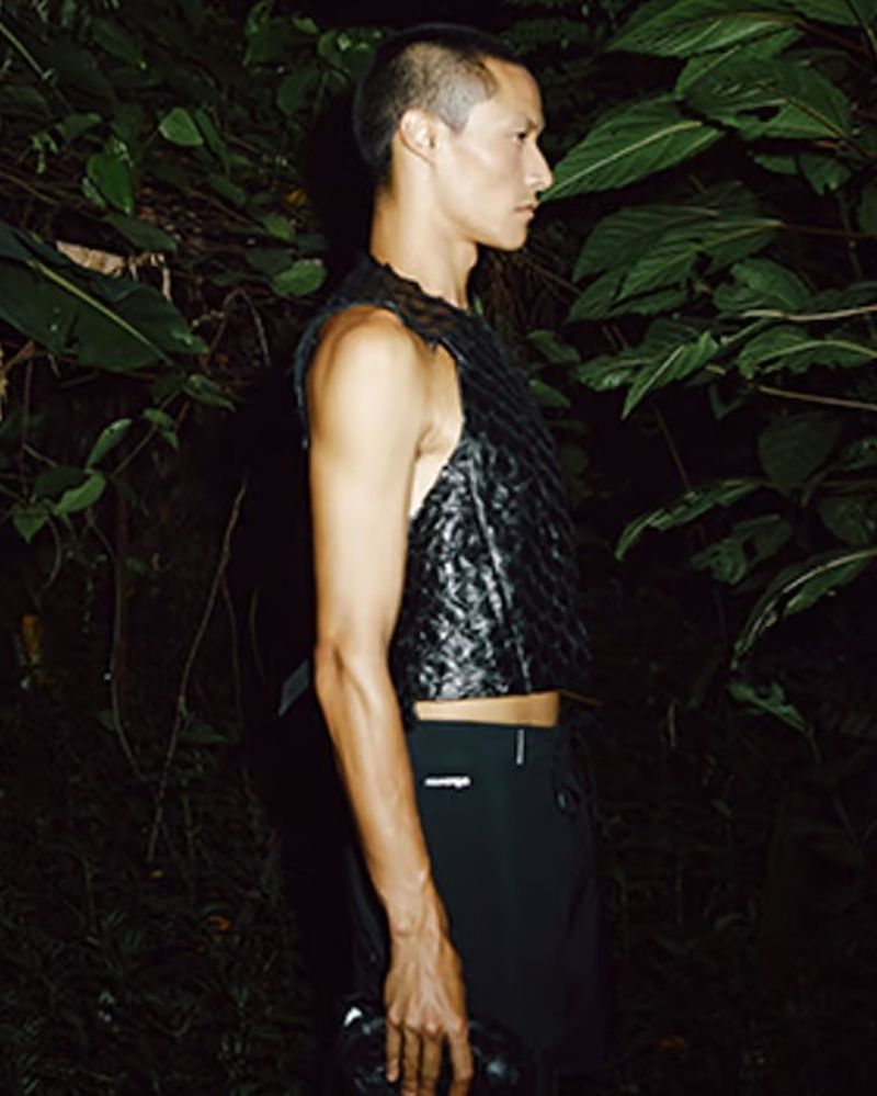 Profile view of an adult male wearing a textured black top and pants, standing amidst dense, dark green foliage, creating a dramatic contrast.