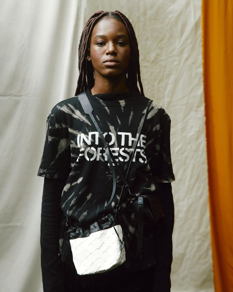 A young Black woman wearing a tie-dye shirt with a shoulder bag poses in front of draped fabric, illuminated by soft, diffused lighting.