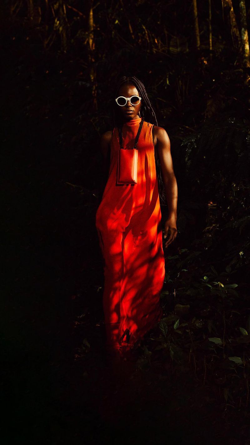 Woman in an orange dress and white sunglasses poses in a dimly lit forest with sun dappling her dress and face.