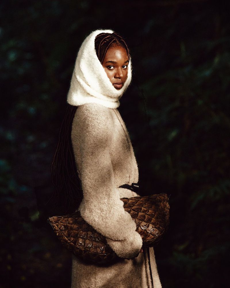 Portrait of a Black woman wearing a wool headscarf and long coat, holding a woven bag, standing against a dark green foliage background.