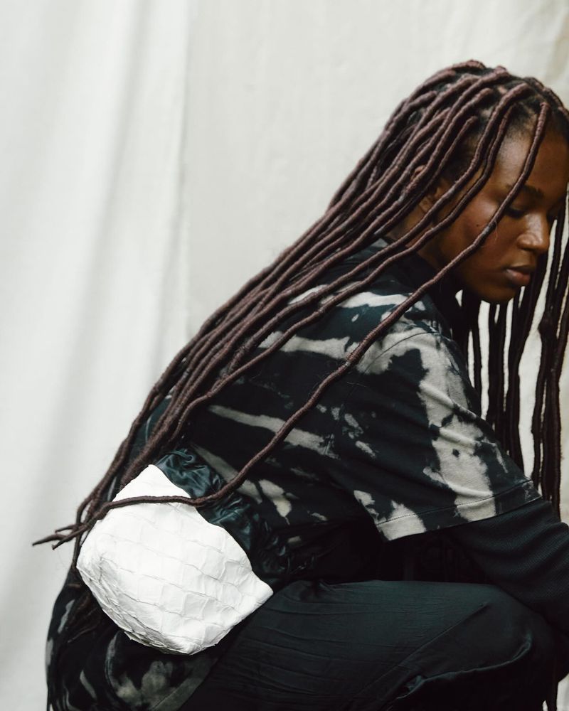 Fashion shot of a young adult woman in black tie-dye, crouched indoors with textured brown locs, wearing a white patterned fanny pack.