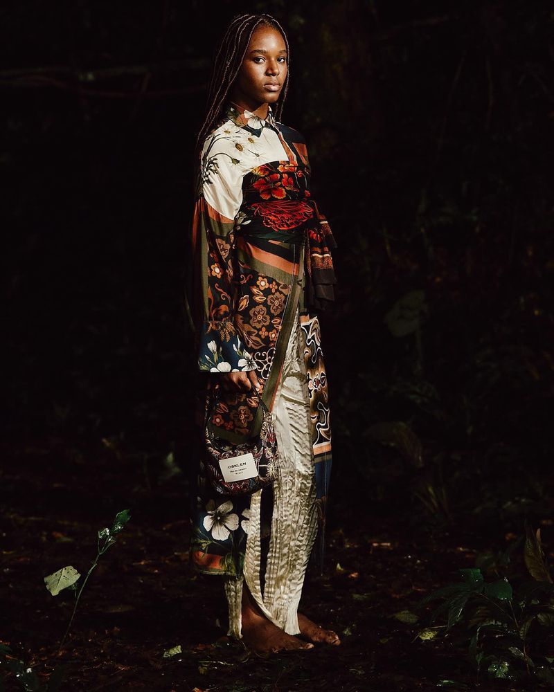 An adult female model with braided hair is posing while standing outside in a long patterned coat with dark background and greenery.