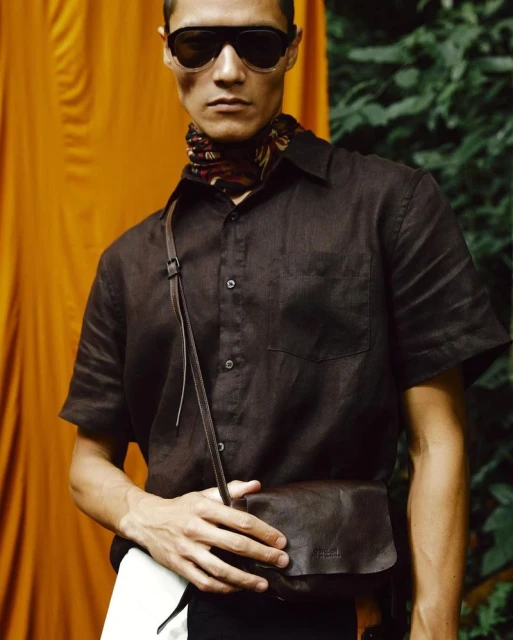 An adult male with sunglasses and a brown shirt is holding a brown leather shoulder bag in front of an orange and green background.