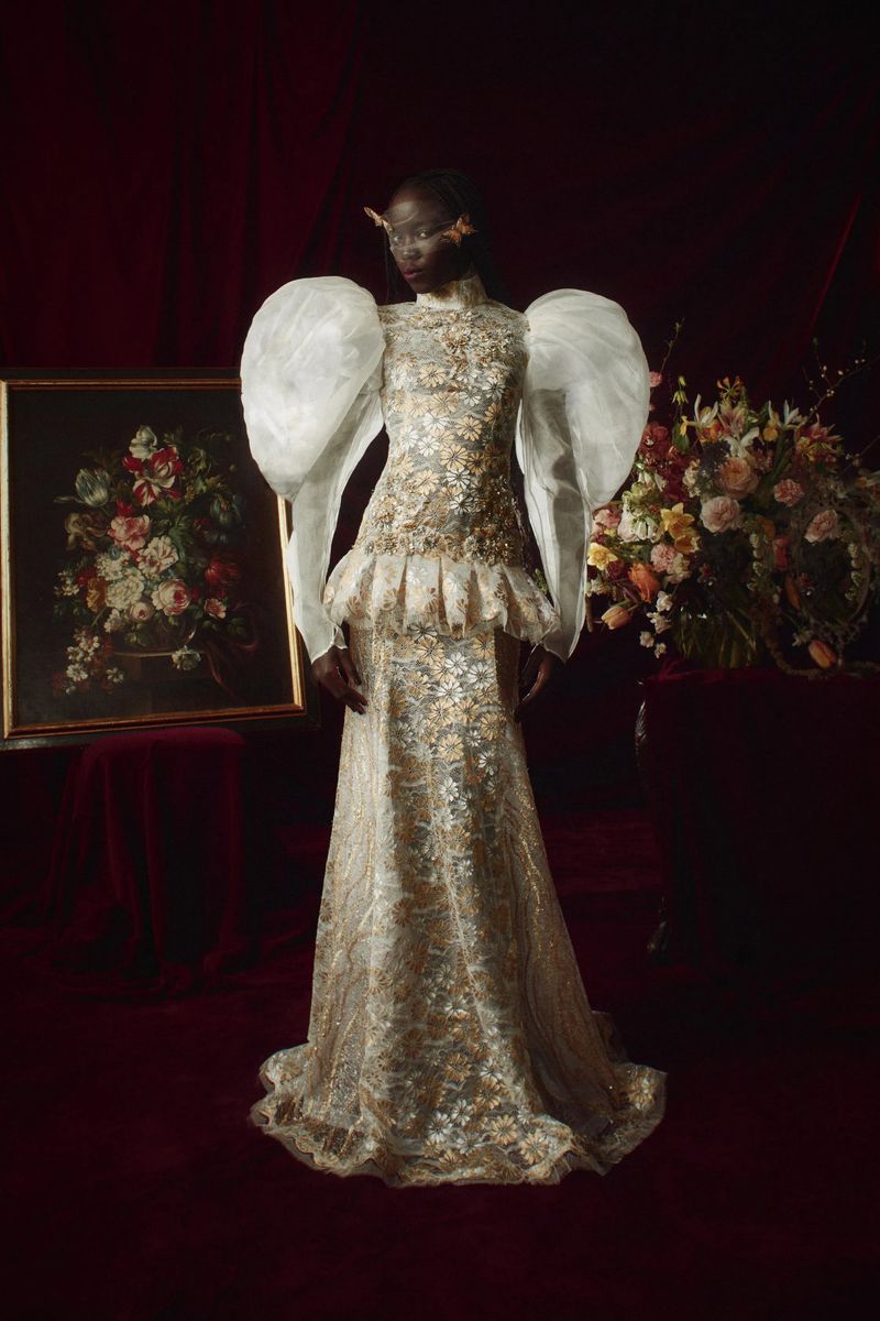 An adult woman poses in a golden gown with large puffed sleeves in front of a dark red curtain and floral arrangement.