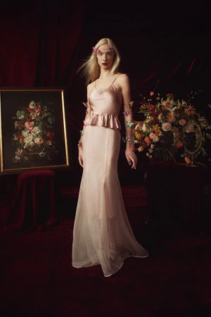 A slender woman with blonde hair stands in a formal gown near an old painting and flowers in a dimly lit room with a red floor.