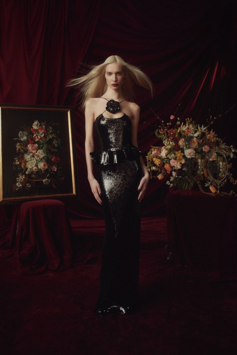 Elegant woman in a black, floor-length gown poses in a dimly lit room with a painting and flowers, showcasing a blend of classic and modern style.