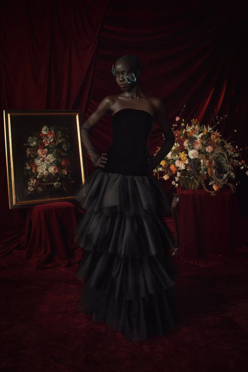 Woman in black gown posing in front of painting and flowers, illuminated by soft light against a rich, dark red velvet backdrop.