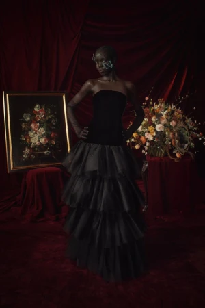Woman in black gown posing in front of painting and flowers, illuminated by soft light against a rich, dark red velvet backdrop.