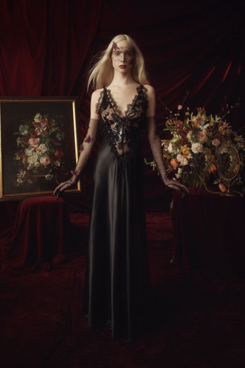 A fair-skinned model in a black gown poses indoors next to a floral painting, with a large floral arrangement in the background.