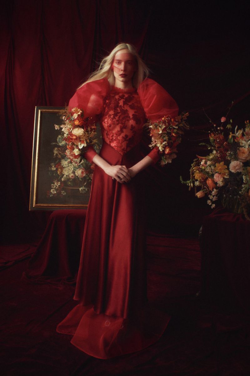 Fashion portrait of a blonde woman in a red dress with floral sleeves standing indoors with a painting and floral arrangement.