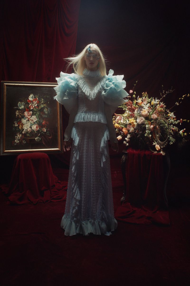 Adult female in a light blue ruffled dress standing in front of a floral painting and a floral arrangement in a dark room.