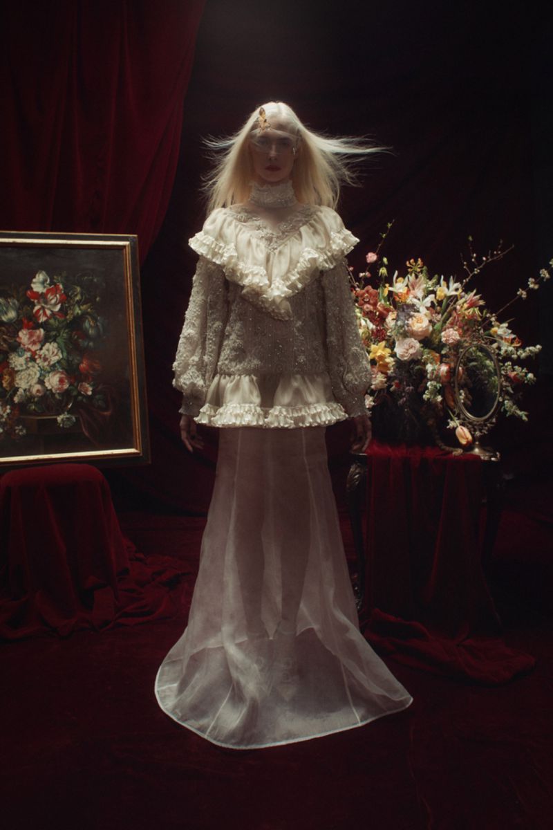 A fashion shot featuring a model in a white vintage-style dress with lace details, standing in front of a floral arrangement and framed painting.