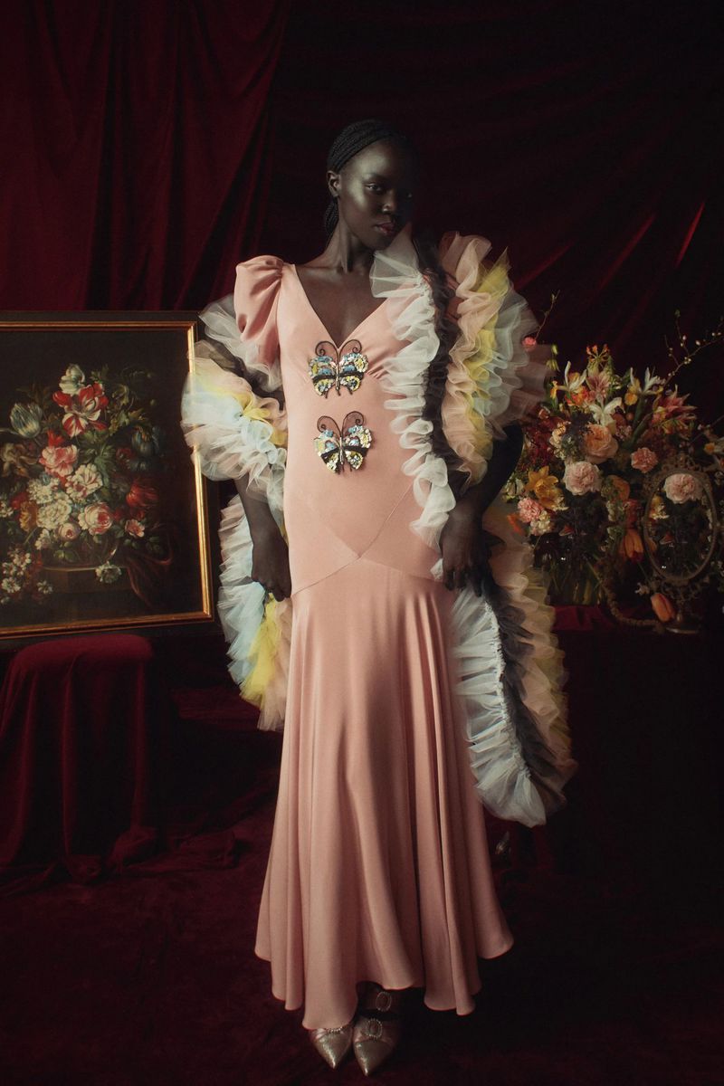 A dark-skinned woman in a blush gown with butterfly embellishments, posing in front of a painting and floral arrangement against a burgundy curtain.