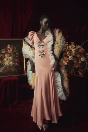 A dark-skinned woman in a blush gown with butterfly embellishments, posing in front of a painting and floral arrangement against a burgundy curtain.