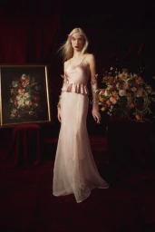 Model in pale pink silk slip dress with ruffled peplum hem standing against dark draped red velvet backdrop with floral arrangements