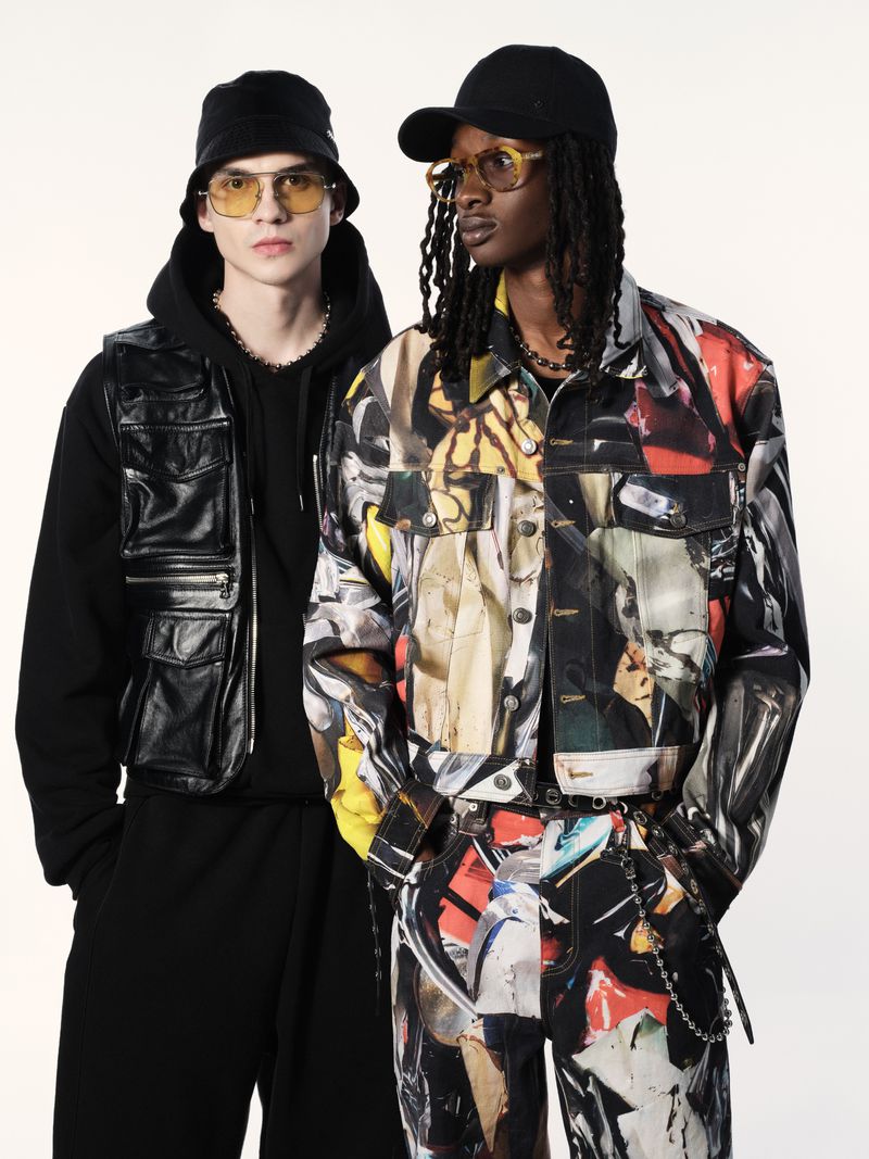 Two adult male fashion models in coordinated abstract print jackets and pants, wearing hats and yellow-tinted glasses, against a white backdrop.