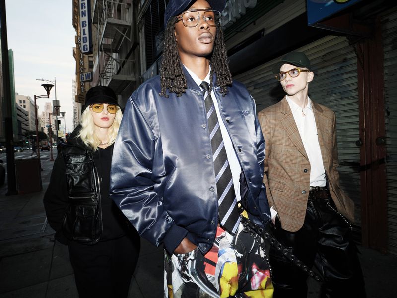 Three people pose on a city street, wearing hats and jackets. One wears a satin bomber jacket and patterned pants, while buildings line the background.