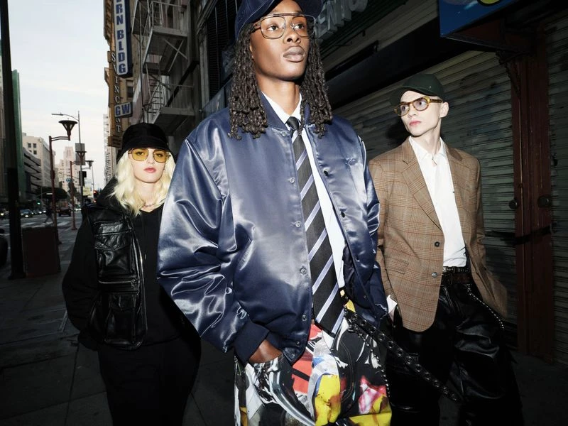 Three people pose on a city street, wearing hats and jackets. One wears a satin bomber jacket and patterned pants, while buildings line the background.