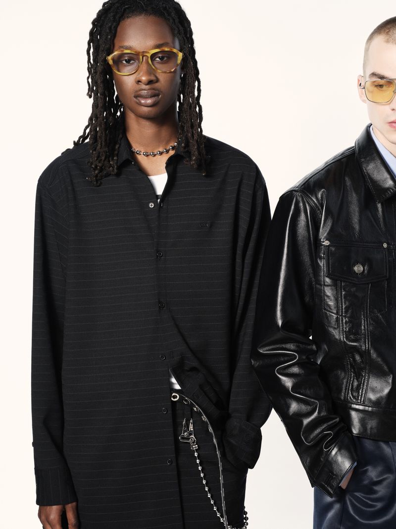 Two fashion models in a studio setting. One wears a black pinstripe shirt and the other a shiny black leather jacket and blue pants.