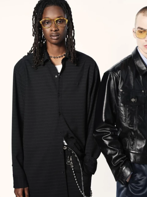 Two fashion models in a studio setting. One wears a black pinstripe shirt and the other a shiny black leather jacket and blue pants.