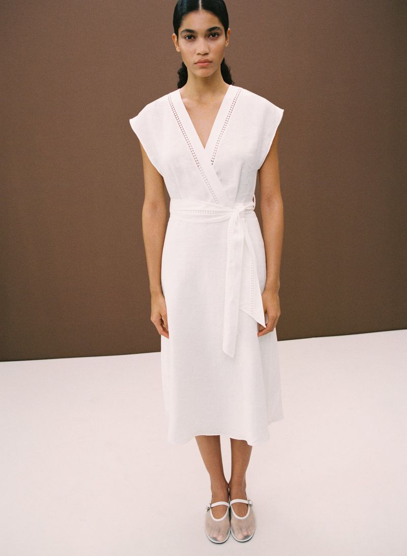 A woman in a white wrap dress with a tie waist stands in a studio with a brown backdrop, wearing Mary Jane shoes.