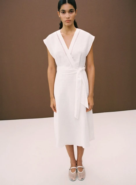 A woman in a white wrap dress with a tie waist stands in a studio with a brown backdrop, wearing Mary Jane shoes.