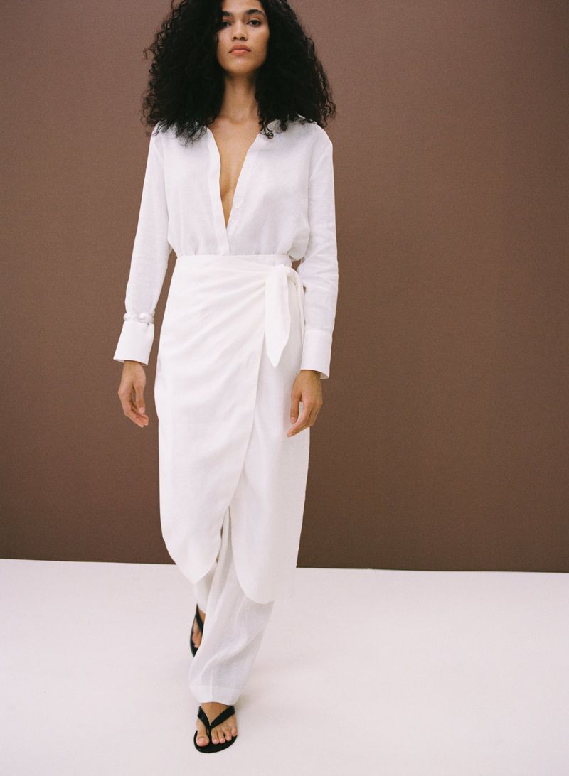 A woman with dark curly hair models a white long-sleeved shirt and wrap skirt with black sandals in a studio setting on a white floor.