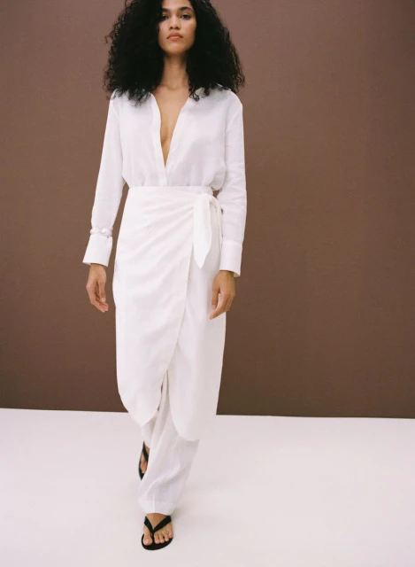A woman with dark curly hair models a white long-sleeved shirt and wrap skirt with black sandals in a studio setting on a white floor.