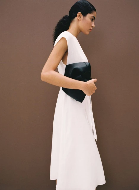 Woman in white dress holds black handbag against a brown background in a fashion setting, with side view and elegant pose.