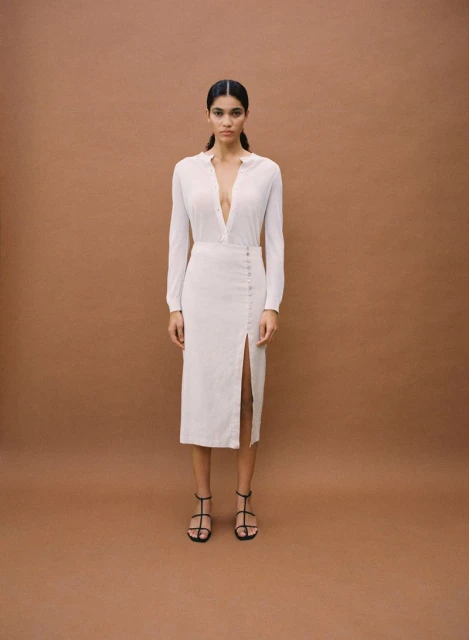 Fashion shot of a woman wearing a long-sleeve top and midi skirt with a slit, paired with strappy heels against a solid brown background.