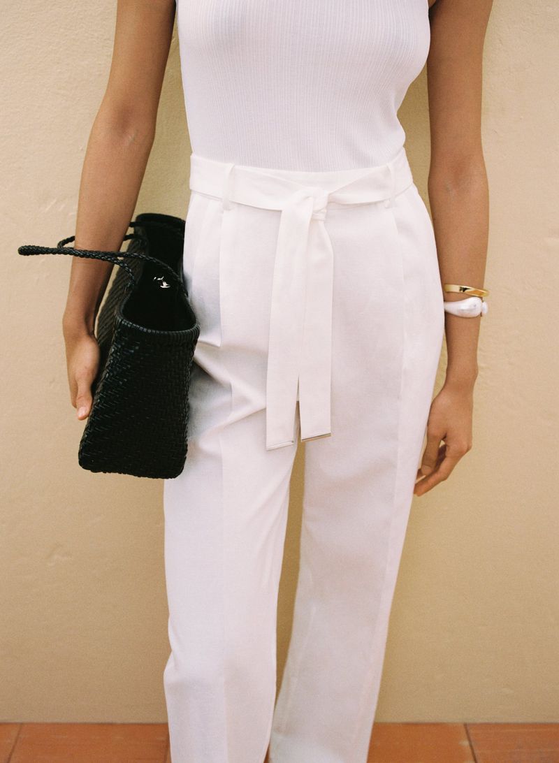 A woman models white belted pants and a white top against a neutral background, holding a black woven bag, showcasing minimalist fashion.
