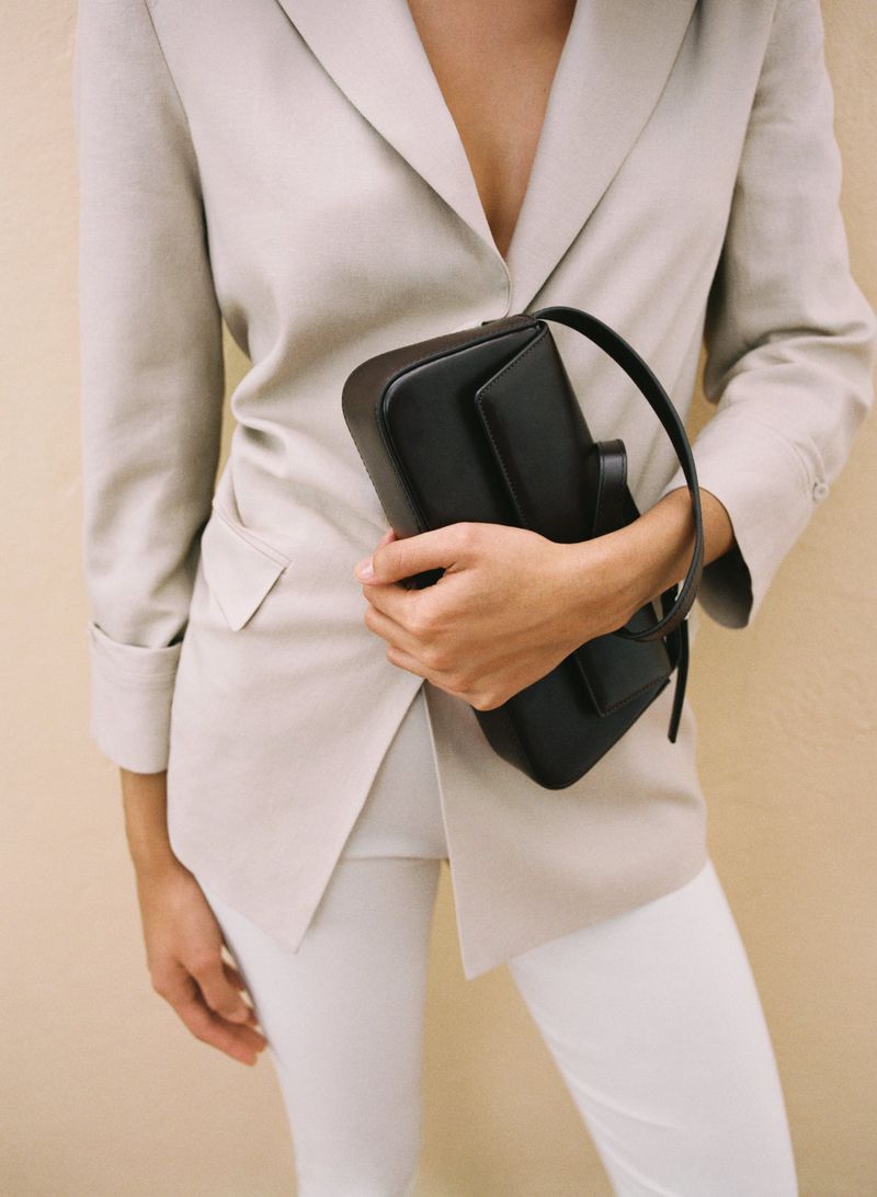 Woman in beige suit jacket and white pants holding black leather purse in indoor setting against tan wall with studio lighting.