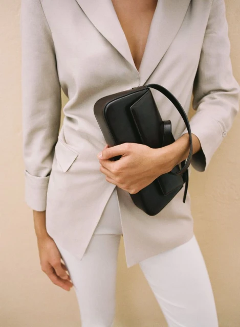 Woman in beige suit jacket and white pants holding black leather purse in indoor setting against tan wall with studio lighting.
