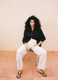 Model Raynara Negrine seated on a chair wearing a black shirt and white linen trousers for Mango Linen Special