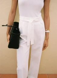 Model wearing white linen trousers with a belted waist and white top holding a black handbag.