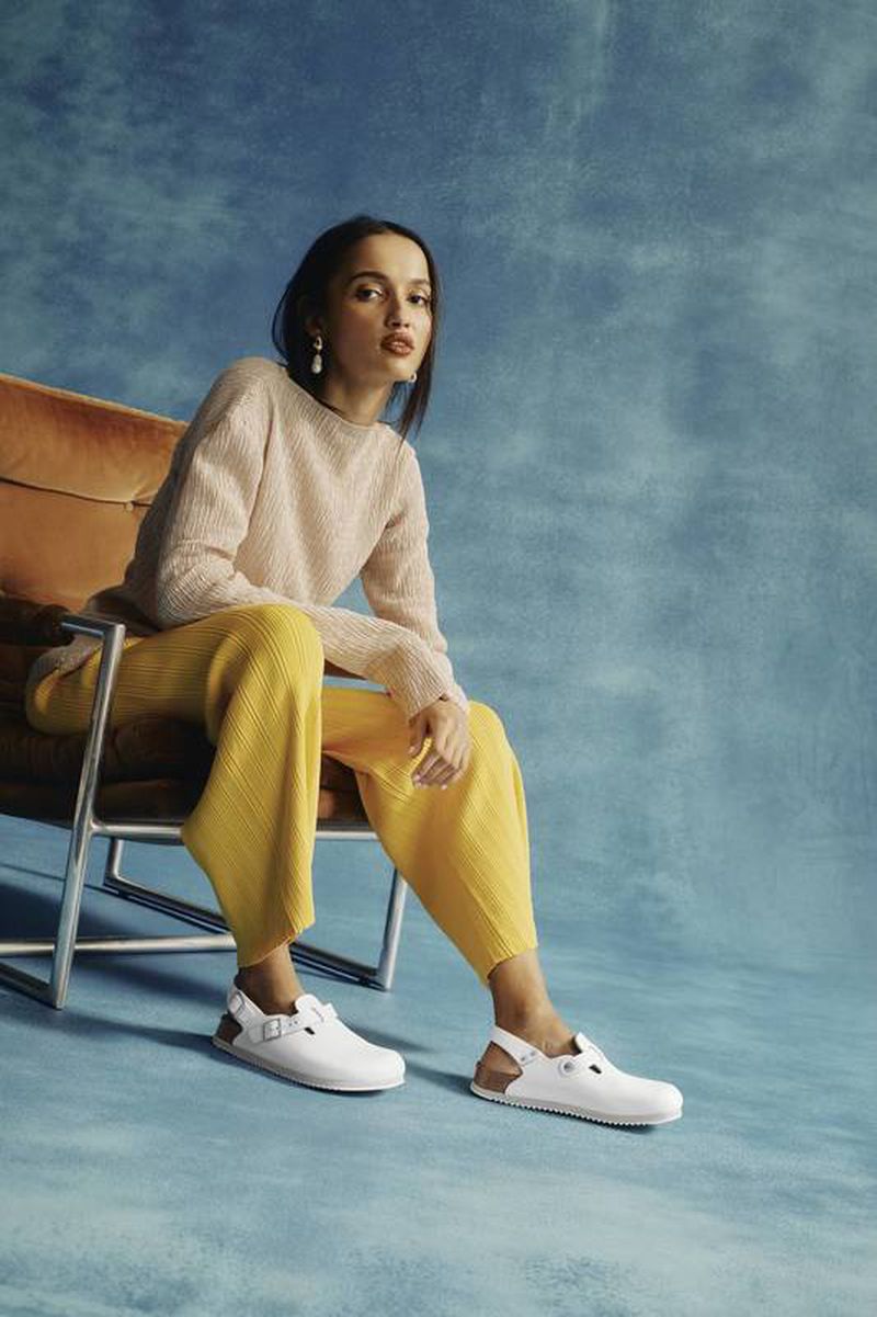 A woman poses in an apricot sweater and yellow pleated pants, seated in a chair against a blue textured background, showcasing white shoes.