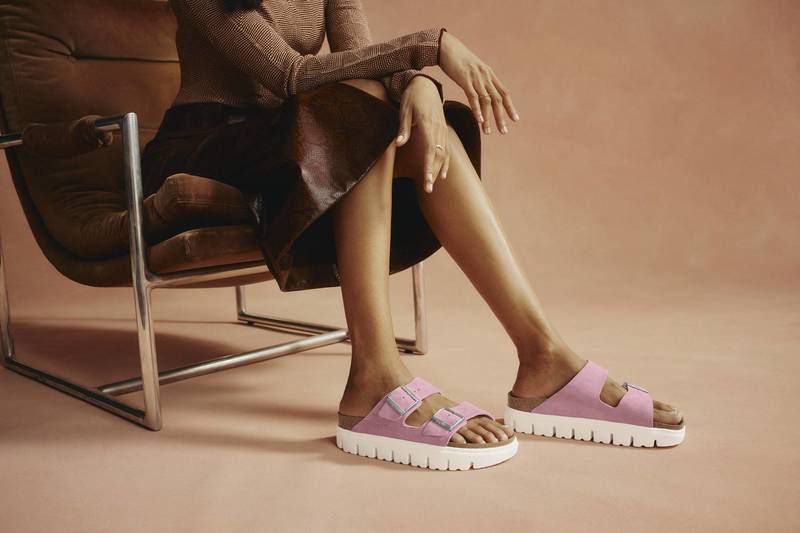 A woman sits in a modern chair, showcasing pink platform sandals with white soles against a light pink backdrop in a studio setting.
