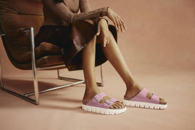 A woman sits in a modern chair, showcasing pink platform sandals with white soles against a light pink backdrop in a studio setting.