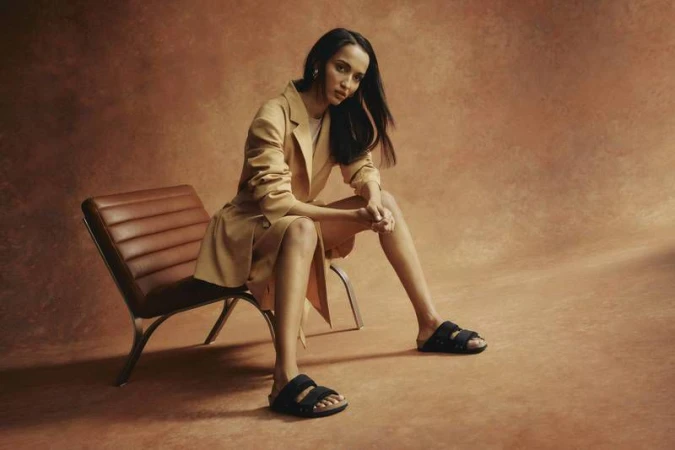Model wearing a tan blazer and black sandals seated on a leather chair against a textured warm background