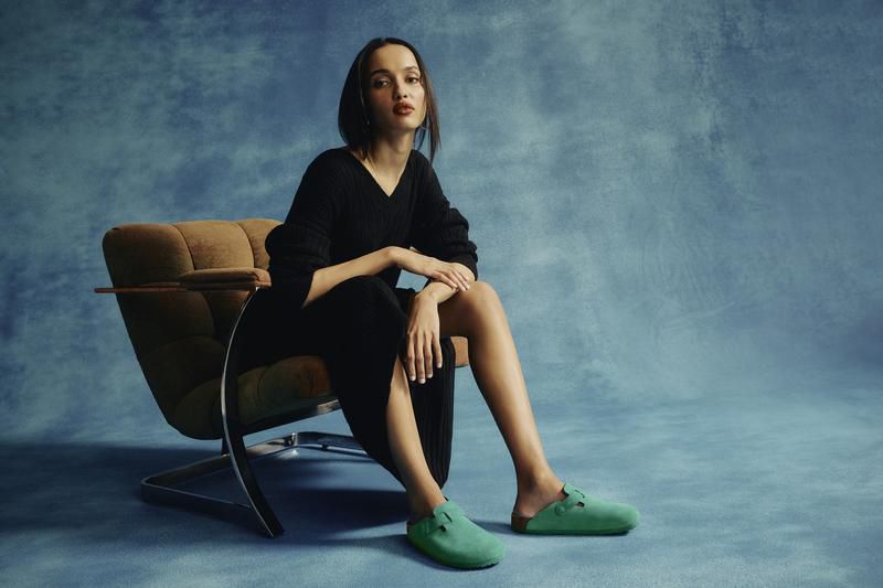 Woman in black dress poses in a brown chair against a blue backdrop, wearing bright green clogs. Fashion editorial style.