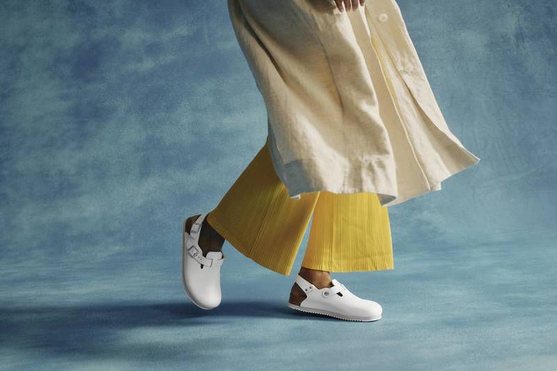 White clogs are featured in a fashion shot of a woman wearing yellow pants and a light jacket against a light blue backdrop.
