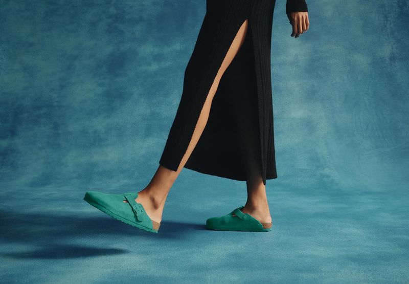 Fashionable green mules are worn by a woman in a long black dress against a mottled blue backdrop in a studio setting.