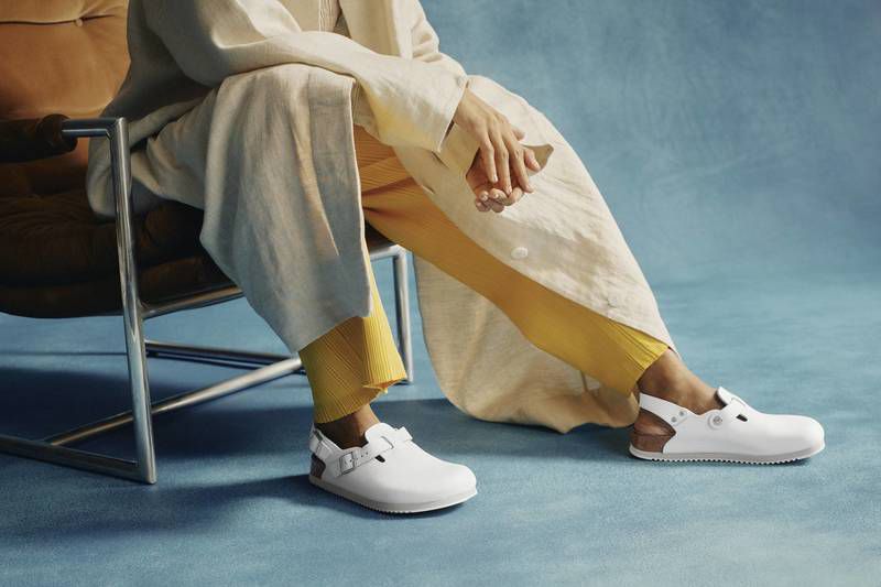 Person sits in a chair wearing white shoes, yellow pants, and a beige coat against a blue backdrop. Casual fashion and style.