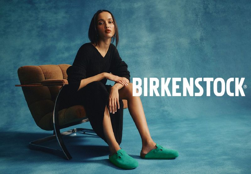 Brunette woman in black dress sits in chair, wearing green Birkenstock clogs against blue mottled background for fashion advertising.