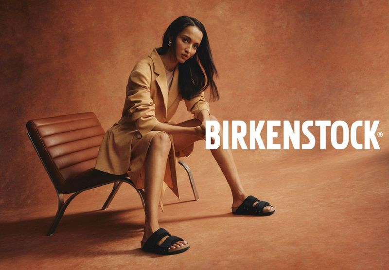 Woman wearing a light brown jacket and black Birkenstock sandals, sitting on a chair in a studio with a brown backdrop.
