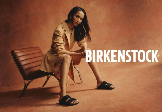 Woman wearing a light brown jacket and black Birkenstock sandals, sitting on a chair in a studio with a brown backdrop.
