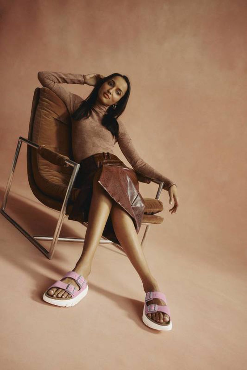 A woman sits in a mid-century modern chair wearing a long-sleeved top, leather skirt, and platform sandals against a peach background.