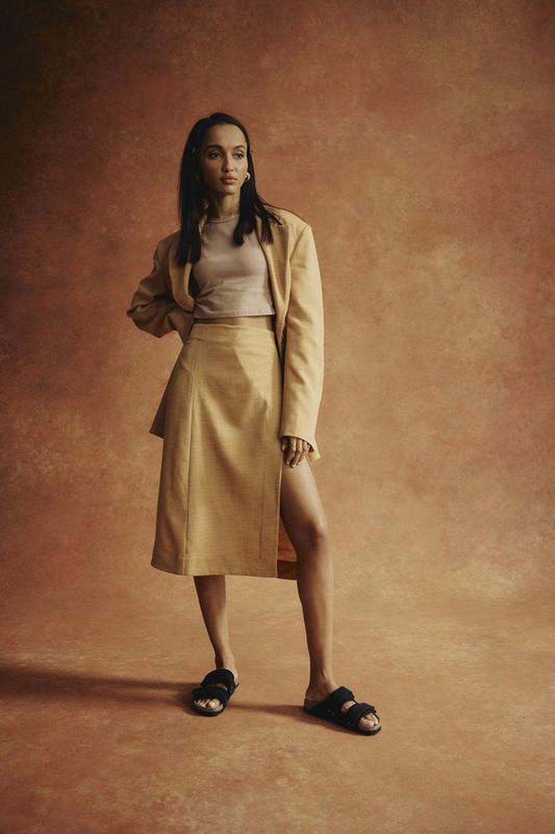 A stylish woman poses in a tan skirt suit and furry black sandals against a textured rust-colored background in a fashion studio setting.