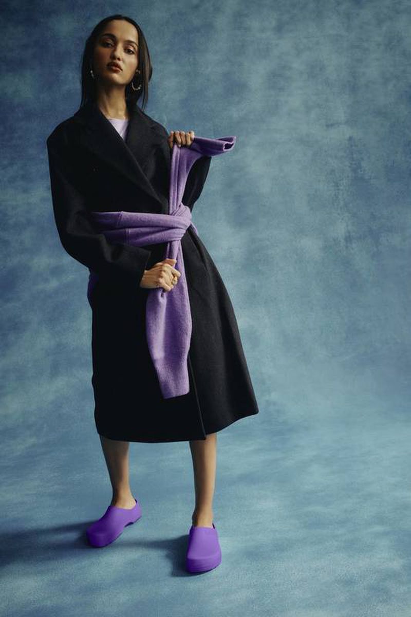 A woman in a black coat and purple shoes poses in front of a textured teal background, holding a purple sweater around her neck.
