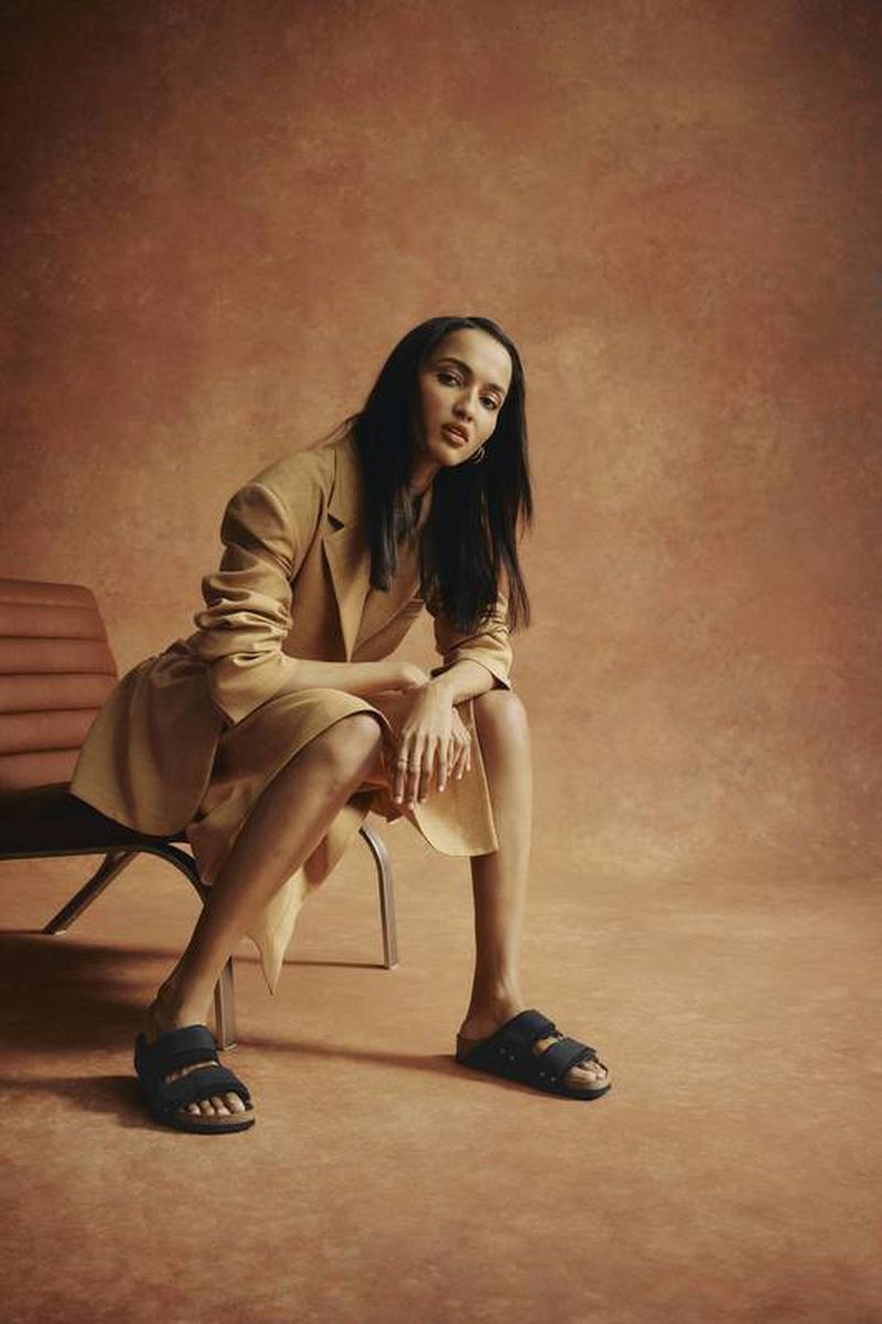 A woman in a tan suit poses seated on a chair in a studio. She wears dark sandals and has long hair. The background is a mottled tan.